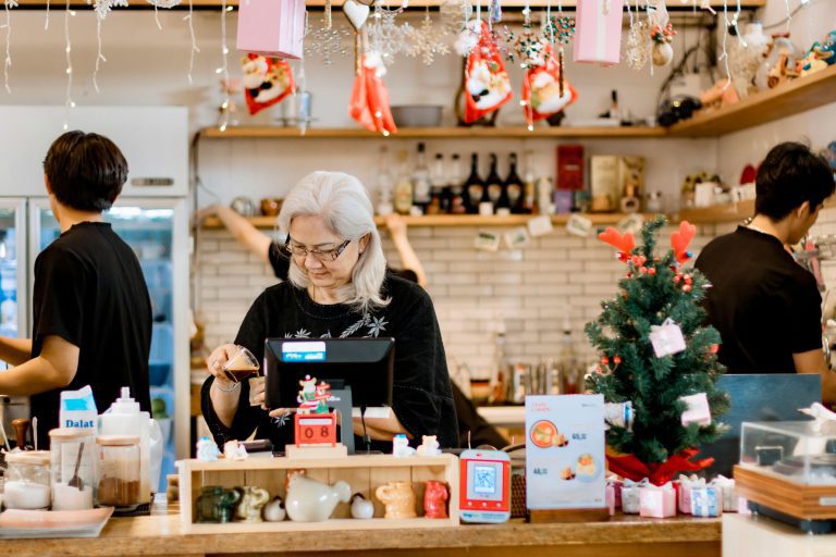 A shopkeeper at the checkout counter of a small retail shop decorated for the holidays