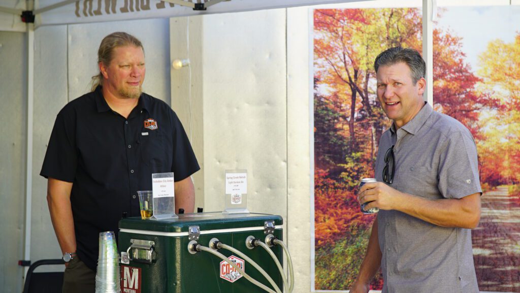 Two men chatting beside a beverage dispenser at an outdoor community event, with an autumn forest banner behind them