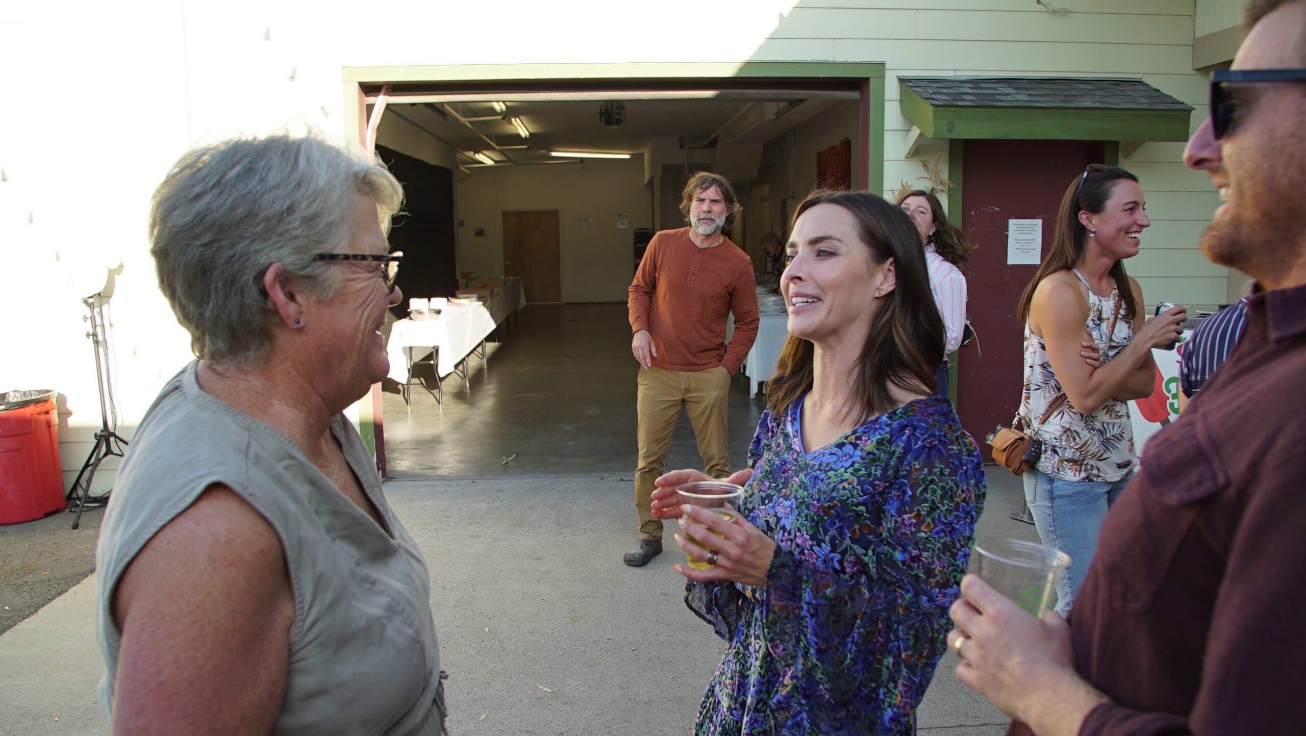 Guests chatting with drinks at an outdoor Mission West event near an open warehouse door at golden hour