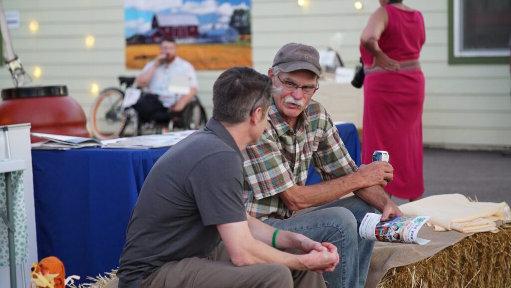 Two guests in conversation on hay bales at the Harvest on Main outdoor dinner, with string lights and a barn backdrop