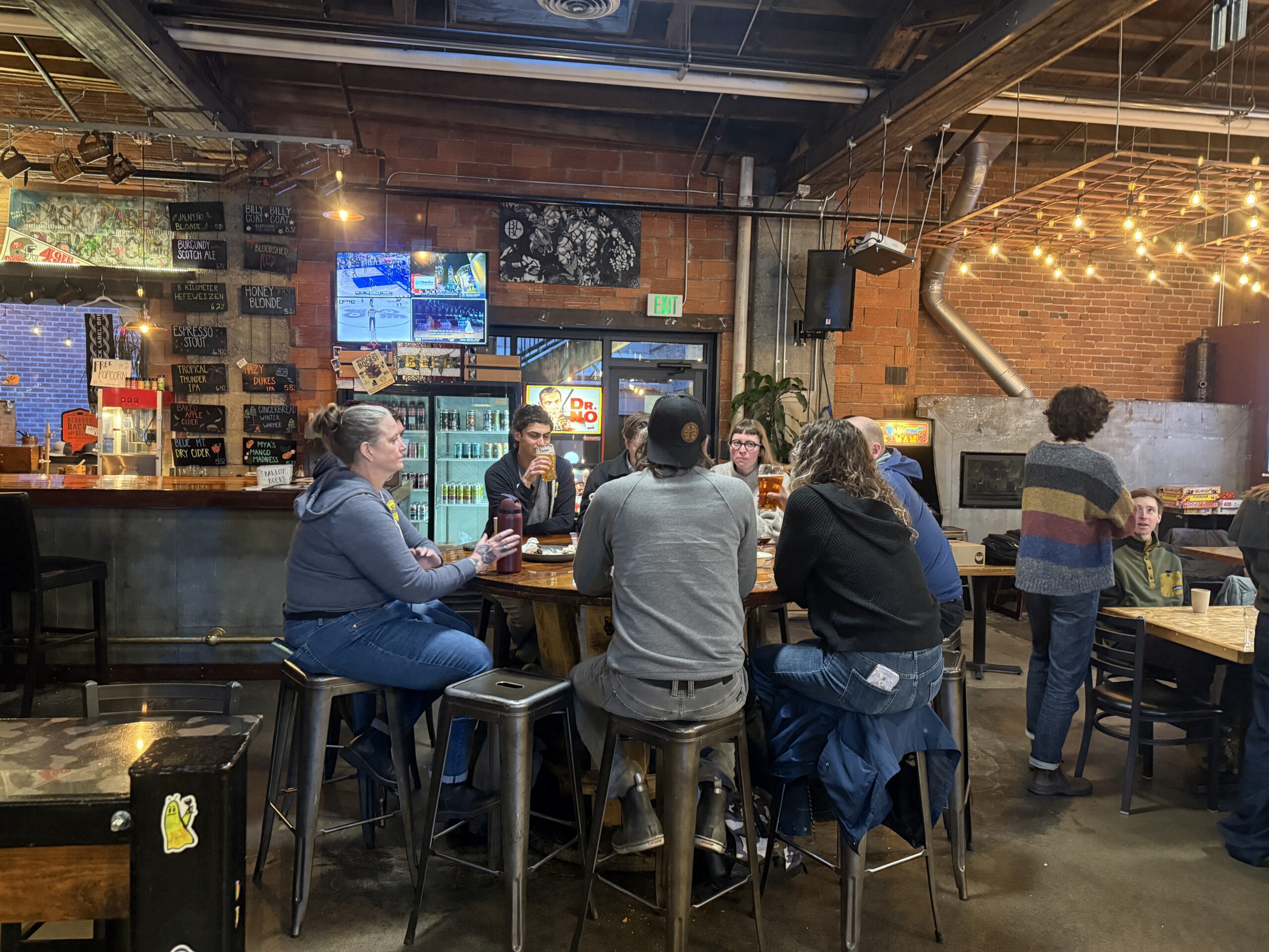 Patrons gathered at high-top tables inside a brewery taproom with brick walls and string lights