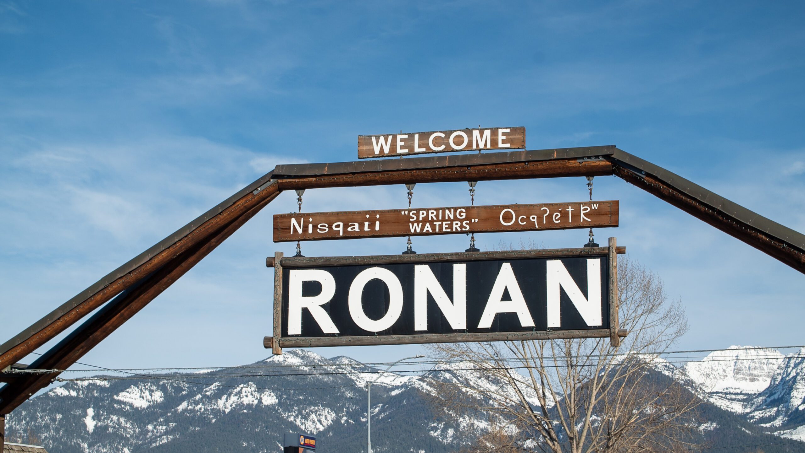 Welcome to Ronan sign with the Salish name Nisqaii (Spring Waters) and the snow-capped Mission Mountains behind